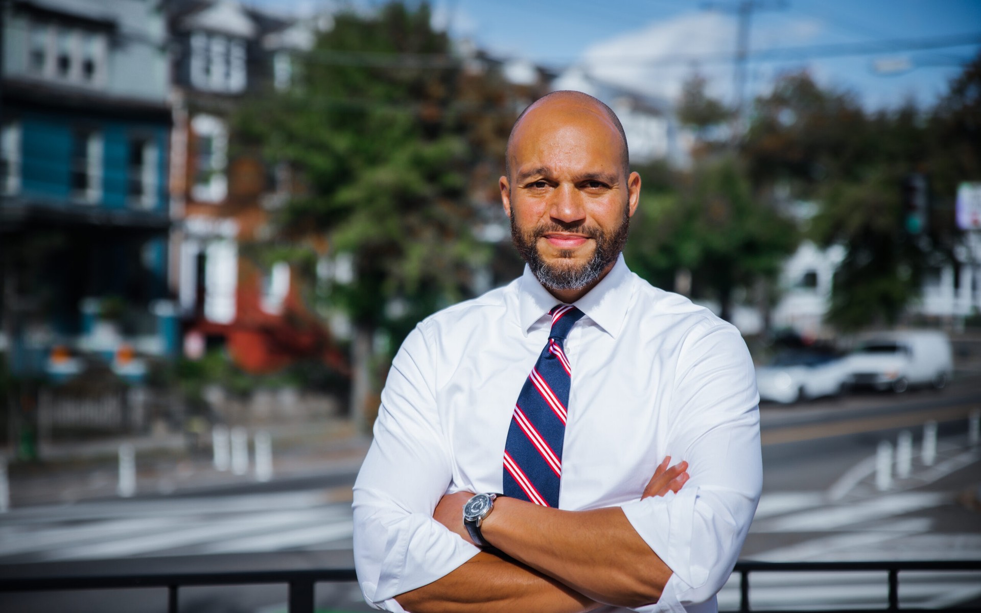 D.C. Councilmember Robert White, in a white shirt and striped tie, standing outdoors with his arms crossed.