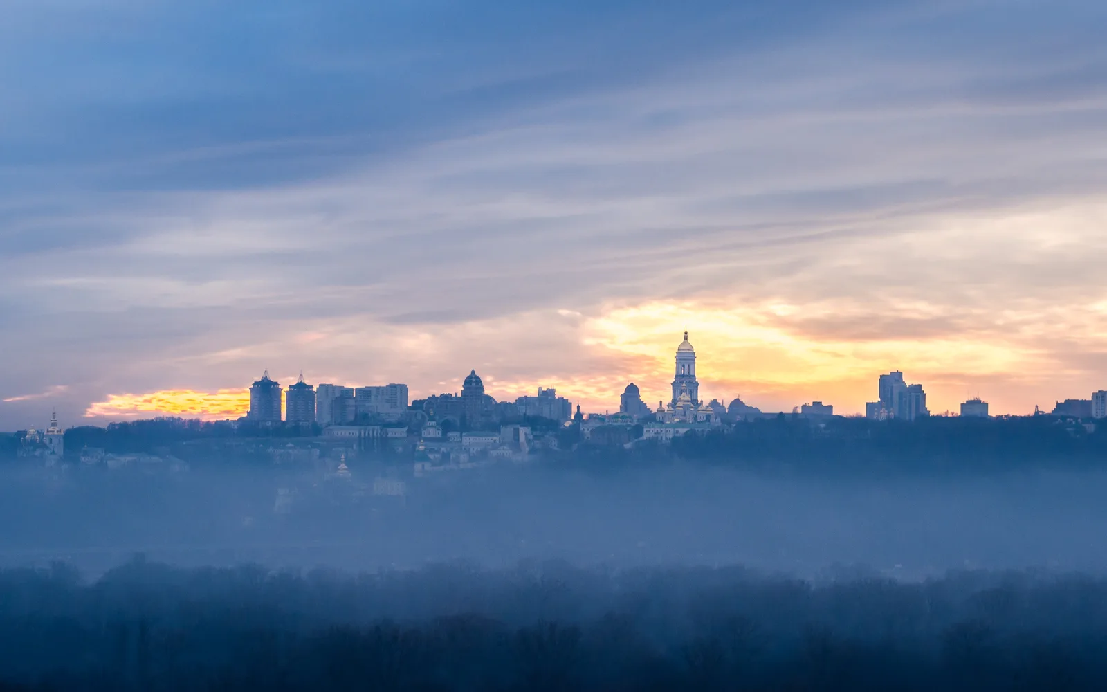 The Kyiv skyline at dawn, with the bell tower of the Kyiv Pechersk Lavra rising above mist over the Dnipro River