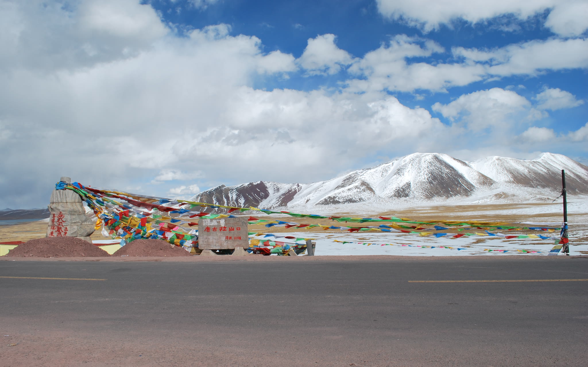Prayer flags strung across a high mountain pass in Nagqu, Tibet, with snow-covered peaks behind.